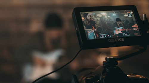 image of a dslr camera filming a man playing guitar and a woman watching, the subjects are out of focus in the background but visible on the monitor of the camera