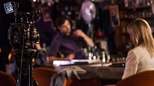 A set photo of a bar scene, figures stand out-of-focus at a bar in the background and a camera in focus in the foreground filming them