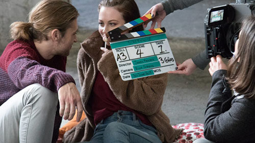 A photo of two actors a man and a woman gazing lovingly at each other the woman has a lollypop. In front of them is a film camera and a clapper board preparing to slate reading roll A3 scene 4 take 1.